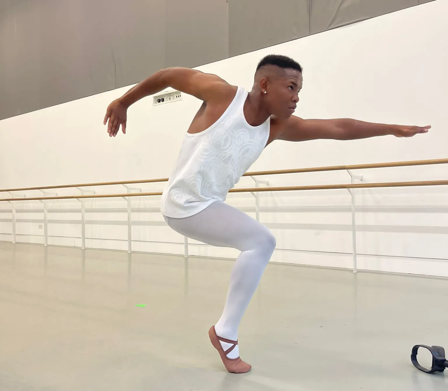 A male ballet dancer with one leg practicing in a studio. He is standing on the toes of one foot, with the other knee bent and foot raised behind him. His arms are extended, one in front and one to the side, creating a dynamic pose. He wears a white sleeveless top with a textured pattern and white tights, and his ballet shoes have crisscrossed straps. To his side, there is a black object on the floor, possibly his crutch.