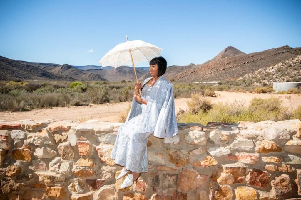 A woman in an elegant, white lace dress with wide sleeves sits on a stone wall. She holds a delicate, lace-trimmed parasol that shades her from the bright sunlight. The backdrop is the serene desert landscape with scattered shrubbery, undulating hills, and a clear blue sky at Aquila Luxury Safari Lodge, Capt Town south Africa. Her poised demeanor suggests a formal or celebratory occasion, contrasting with the rustic, natural surroundings.
