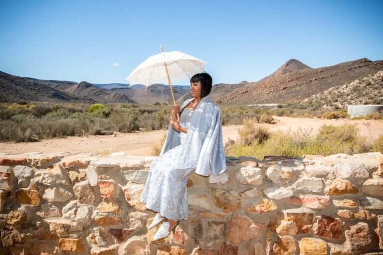 A woman in an elegant, white lace dress with wide sleeves sits on a stone wall. She holds a delicate, lace-trimmed parasol that shades her from the bright sunlight. The backdrop is the serene desert landscape with scattered shrubbery, undulating hills, and a clear blue sky at Aquila Luxury Safari Lodge, Capt Town south Africa. Her poised demeanor suggests a formal or celebratory occasion, contrasting with the rustic, natural surroundings.