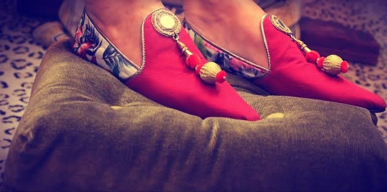A pair of vibrant red traditional pointed shoes with decorative gold pom-poms and embellishments. The shoes have a detailed floral pattern on the heel section. They are worn with red socks and rest on a mustard-colored pillow or cushion with a subtle leopard print in the background, suggesting a cozy and stylish indoor setting.