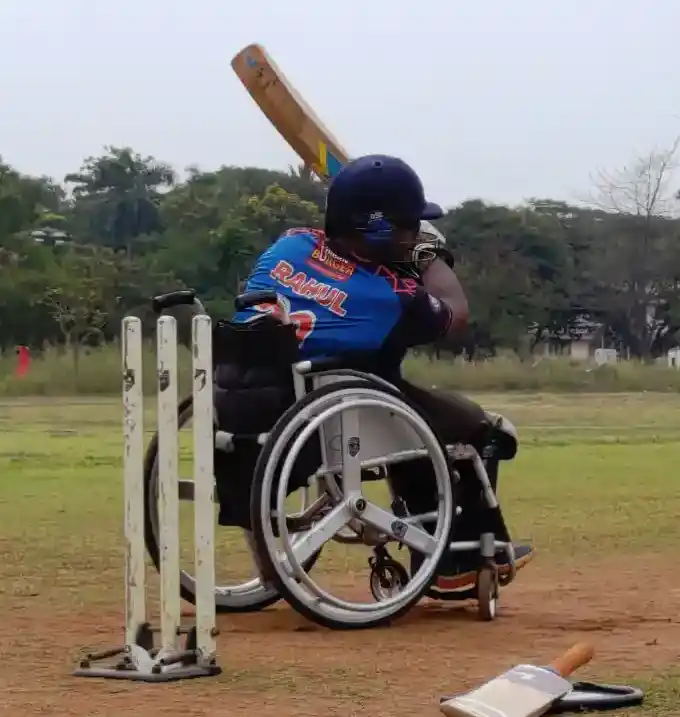 This image shows a wheelchair cricket player on the field. The player is donned in a blue and orange cricket jersey with the name "RAHUL" printed on the back, indicating either the player's name or a sponsored athlete. The player is wearing protective gear, including a helmet and pads, which suggests they are batting. The cricket bat is captured mid-swing, implying that the player has just hit the ball. Three stumps with bails are present, which represent the wicket in cricket. Another bat lies on the ground, suggesting that the player might have changed bats or there was another player present earlier. The background is an open field with trees and a hazy sky, typical of an outdoor cricket field.