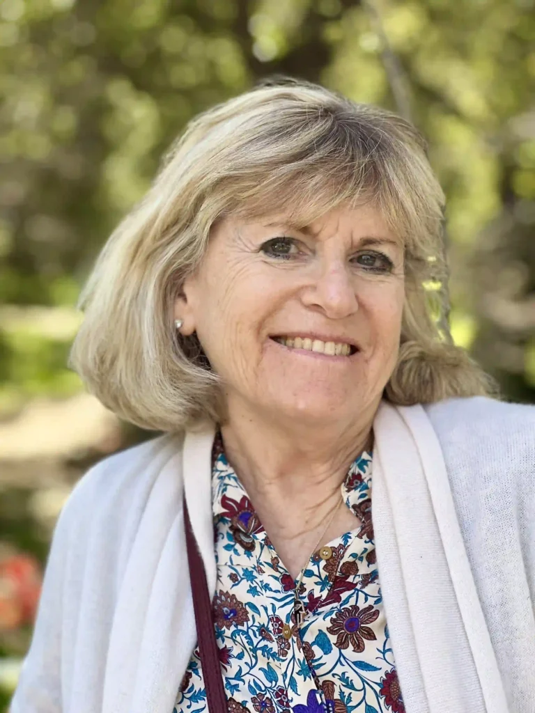 A portrait of a smiling woman with shoulder-length blonde hair. She's wearing a white cardigan over a blouse adorned with a colorful floral pattern, predominantly in shades of blue, with hints of red and yellow. A maroon bag strap crosses over her left shoulder. The background is blurred but appears to be a sunny, verdant outdoor setting.