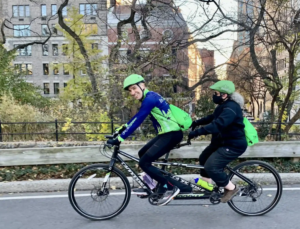 Two people riding a tandem bicycle along a park-side road. The person in the front, smiling at the camera, is wearing a blue jacket with green sleeves, a helmet, and has a water bottle attached to the bike frame. The rider at the back, facing away from the camera, is in black attire with a green helmet and green backpack. The setting is urban, with trees in autumn colors, a black fence, and buildings in the background. There's a sense of motion and recreational activity in an urban environment.