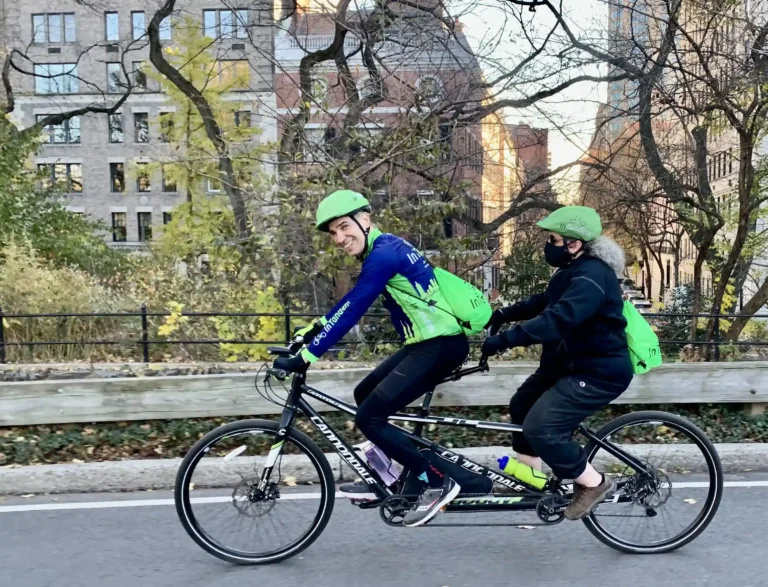 Two people riding a tandem bicycle along a park-side road. The person in the front, smiling at the camera, is wearing a blue jacket with green sleeves, a helmet, and has a water bottle attached to the bike frame. The rider at the back, facing away from the camera, is in black attire with a green helmet and green backpack. The setting is urban, with trees in autumn colors, a black fence, and buildings in the background. There's a sense of motion and recreational activity in an urban environment.