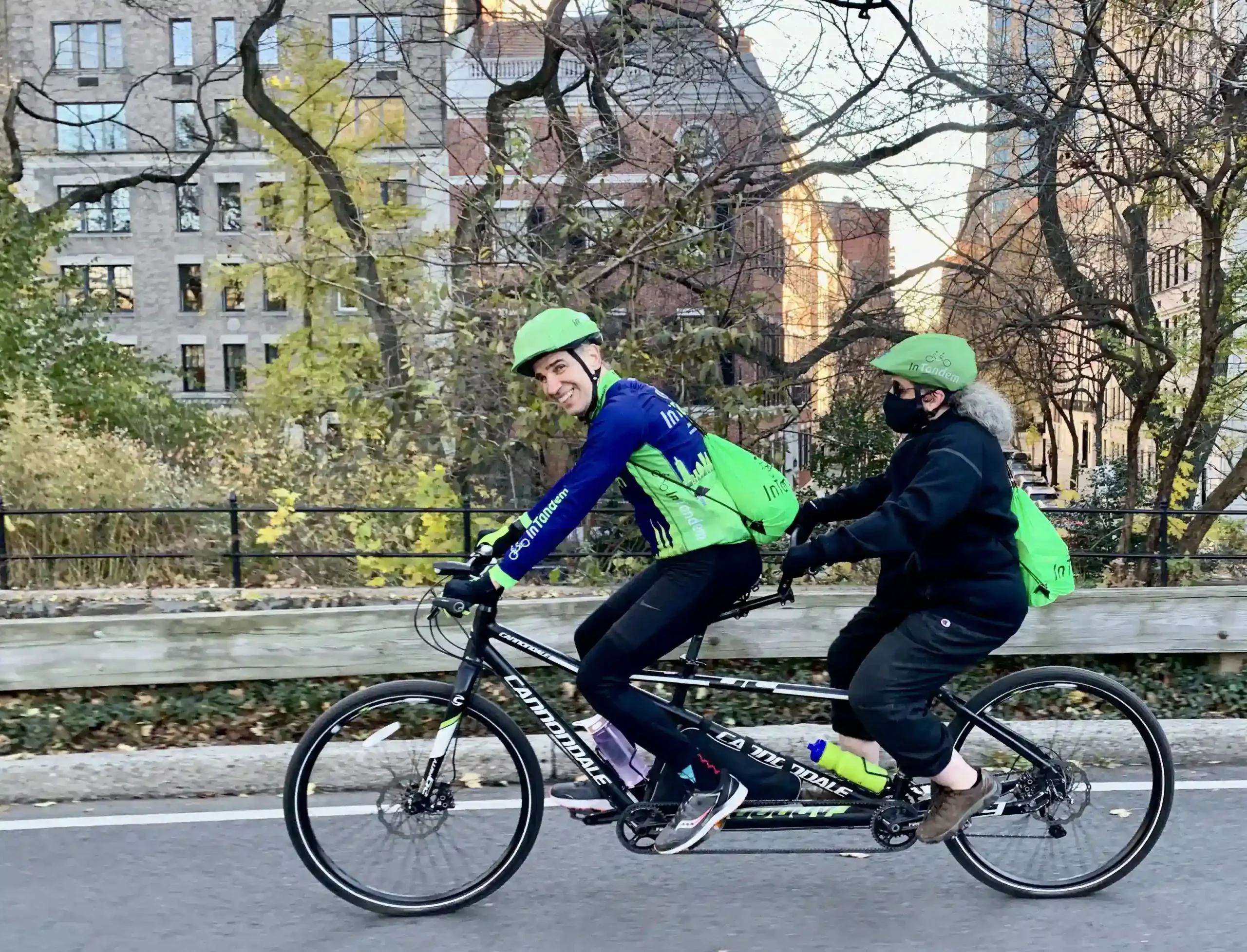 Two people riding a tandem bicycle along a park-side road. The person in the front, smiling at the camera, is wearing a blue jacket with green sleeves, a helmet, and has a water bottle attached to the bike frame. The rider at the back, facing away from the camera, is in black attire with a green helmet and green backpack. The setting is urban, with trees in autumn colors, a black fence, and buildings in the background. There's a sense of motion and recreational activity in an urban environment.