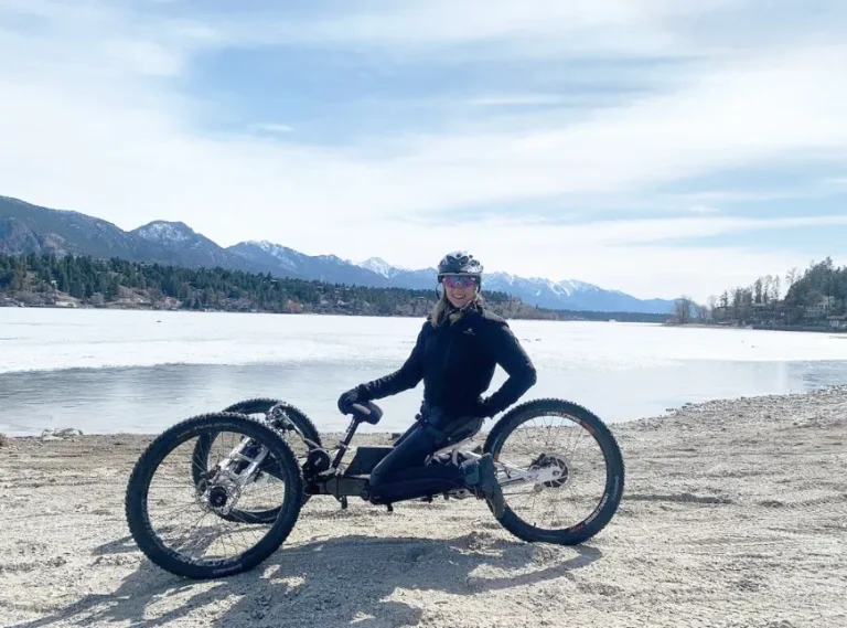 A woman with a joyful expression is seated on a unique three-wheeled bike on a pebbly shore beside a lake. She is dressed in black athletic attire, complete with a helmet, and gloves, ready for an active day out. The serene lake is partially frozen, indicating a cool climate, with a backdrop of majestic mountains under a partly cloudy sky. The natural scenery suggests a tranquil setting ideal for outdoor activities.