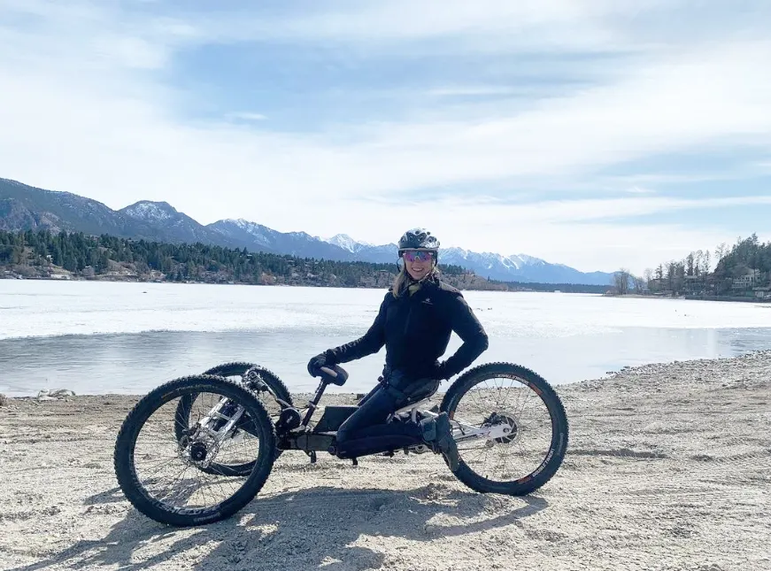 A woman with a joyful expression is seated on a unique three-wheeled bike on a pebbly shore beside a lake. She is dressed in black athletic attire, complete with a helmet, and gloves, ready for an active day out. The serene lake is partially frozen, indicating a cool climate, with a backdrop of majestic mountains under a partly cloudy sky. The natural scenery suggests a tranquil setting ideal for outdoor activities.