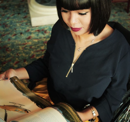 A woman with a stylish bob haircut, reading a large, open book. She is wearing a dark blouse with long sleeves and a distinctive zipper pendant necklace. The setting appears to be a room with a patterned carpet, suggesting a comfortable and elegant ambiance.