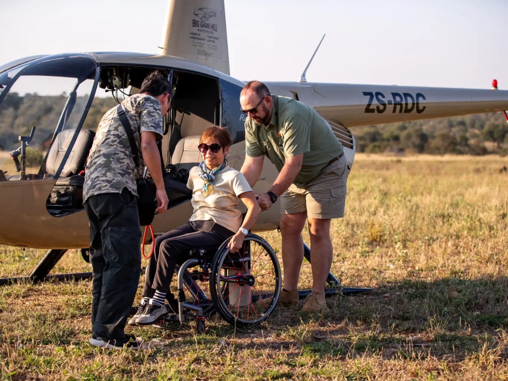 A woman in a wheelchair is being assisted by two men near a small helicopter in a field. The helicopter's tail number, ZS-RDC, suggests it may be registered in South Africa. The woman is wearing sunglasses, a cream-colored shirt, and black trousers. One man, wearing a camouflage shirt and black pants, is on the left, assisting her with something in her lap, possibly a bag or safety harness. The other man, dressed in a green shirt and beige shorts, is on the right, holding the wheelchair steady. The atmosphere appears calm, possibly post-flight, and they are all focused on the task at hand. The field behind them is vast and open, suggesting a rural or safari setting.