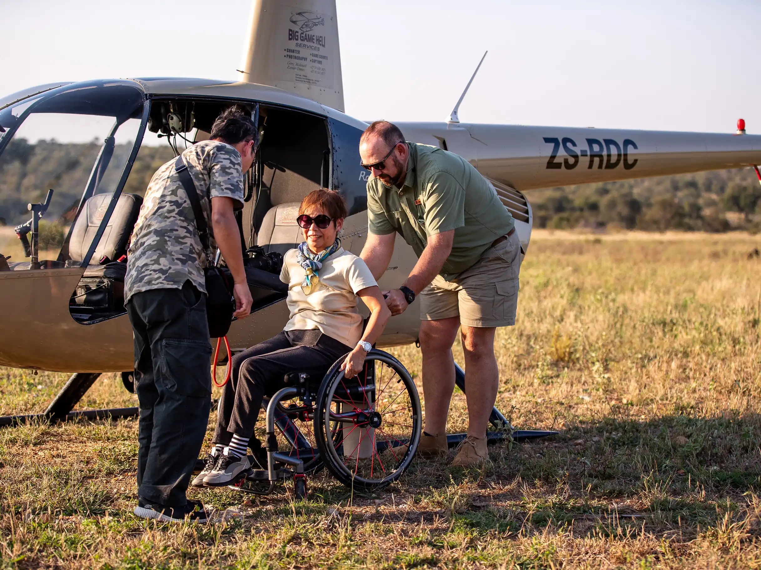 A woman in a wheelchair is being assisted by two men near a small helicopter in a field. The helicopter's tail number, ZS-RDC, suggests it may be registered in South Africa. The woman is wearing sunglasses, a cream-colored shirt, and black trousers. One man, wearing a camouflage shirt and black pants, is on the left, assisting her with something in her lap, possibly a bag or safety harness. The other man, dressed in a green shirt and beige shorts, is on the right, holding the wheelchair steady. The atmosphere appears calm, possibly post-flight, and they are all focused on the task at hand. The field behind them is vast and open, suggesting a rural or safari setting.