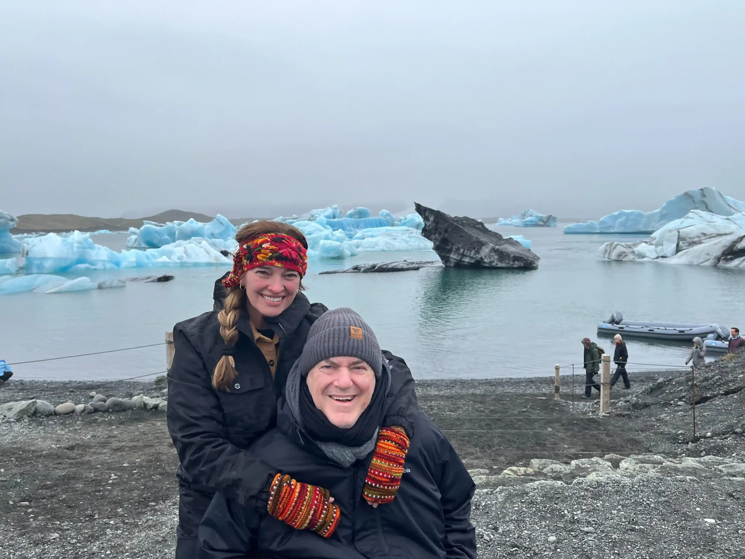 A cheerful couple posing for a photo in a cold, overcast climate. The woman, on the left, has a big smile and is wearing a dark jacket and a colorful headband, her hair styled in a braid. The man is grinning broadly, sporting a beanie and a dark coat with bright, multi-colored wristbands. Behind them, an icy landscape unfolds with multiple blue-white icebergs floating in a calm body of water. To the right, a few people can be seen walking near the shore and an inflatable boat, hinting at a tourist setting likely in a glacial lagoon.