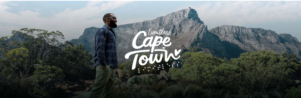 A man stands on the left side of the image, gazing off into the distance. He is stylishly dressed in a blue button-up shirt and olive green pants. The majestic Table Mountain looms in the background, beneath a vast, cloud-streaked sky. The foliage of the Cape Floristic Region is prominent, giving a sense of wild beauty. Overlaid on the image are the words "Limitless Cape Town" in a playful, white script, accentuating the theme of boundless adventure and natural grandeur in this South African city.