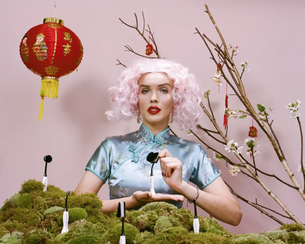 A woman with pink curly hair is dressed in a traditional blue Chinese cheongsam with floral embroidery. She holds an adaptive makeup brush in her right hand and is positioned against a pink backdrop. To her right, a branch with white blossoms and small red lantern decorations extends into the frame. Above her is a traditional red Chinese lantern, and in front of her, there is a patch of moss with several small black objects that appear to be microphone stands with labels. The scene has a whimsical and stylized feel, combining elements of traditional Chinese culture with a modern, colorful aesthetic.