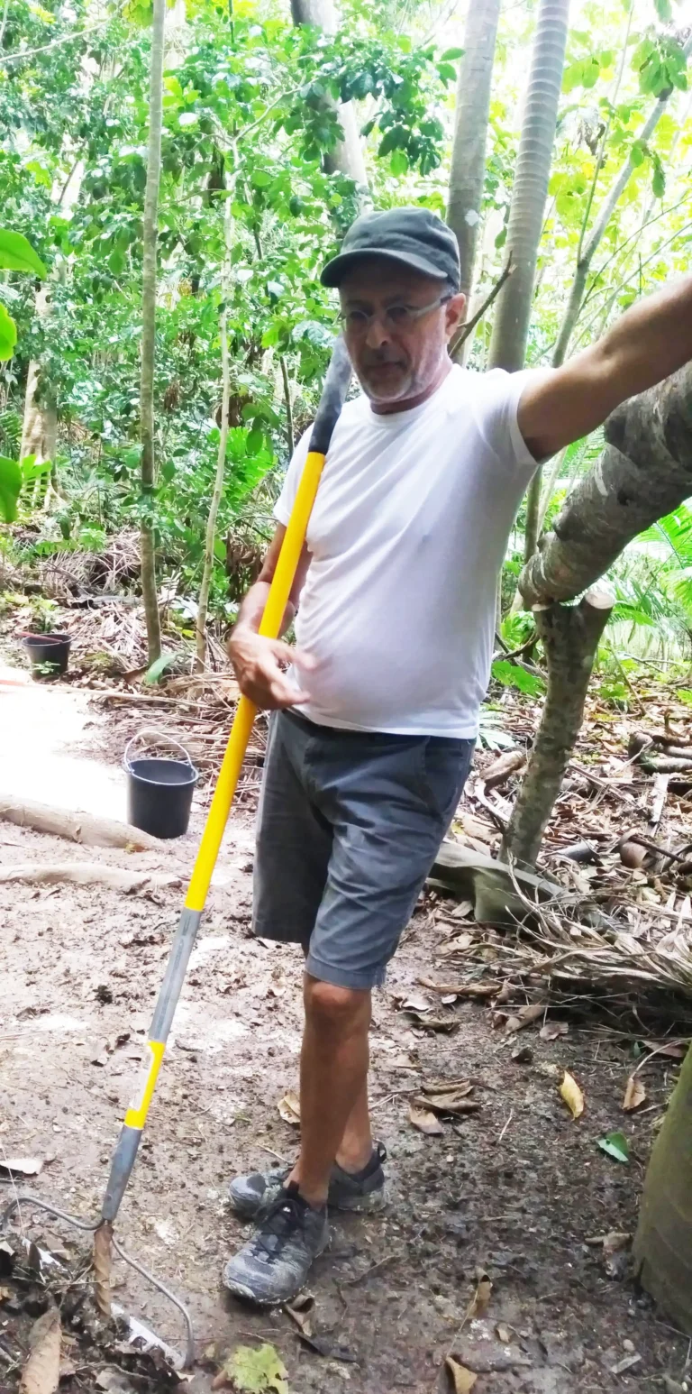 A man wearing a white t-shirt, grey shorts, a black cap, and glasses is standing in a forested area. He is holding a yellow-handled garden tool and leaning against a tree with his right arm. The ground around him is covered with leaves, branches, and some gravel. A black bucket is placed on the ground nearby. The background is filled with lush green foliage, suggesting a dense forest or jungle environment. The man appears to be taking a break from working.