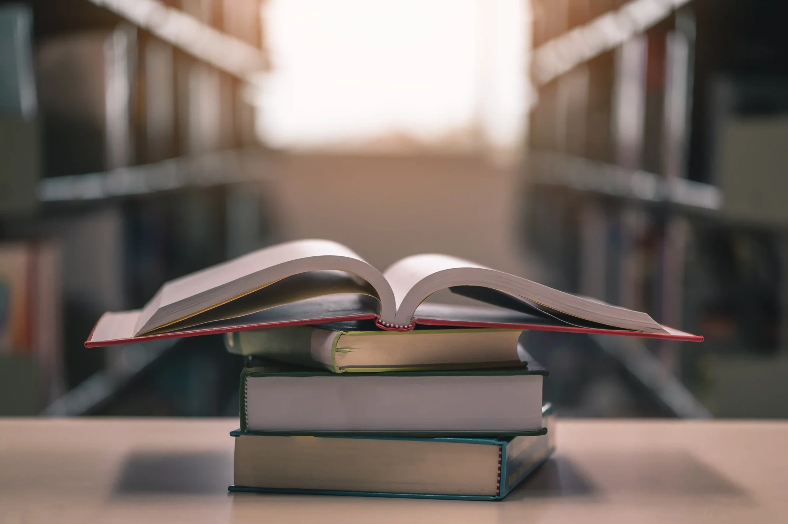 The image shows an open book resting on top of a small stack of closed books. The scene is set in a library or a bookstore, with shelves of books visible in the blurred background. The light filtering in from the background adds a warm, inviting glow to the setting, highlighting the pages of the open book.