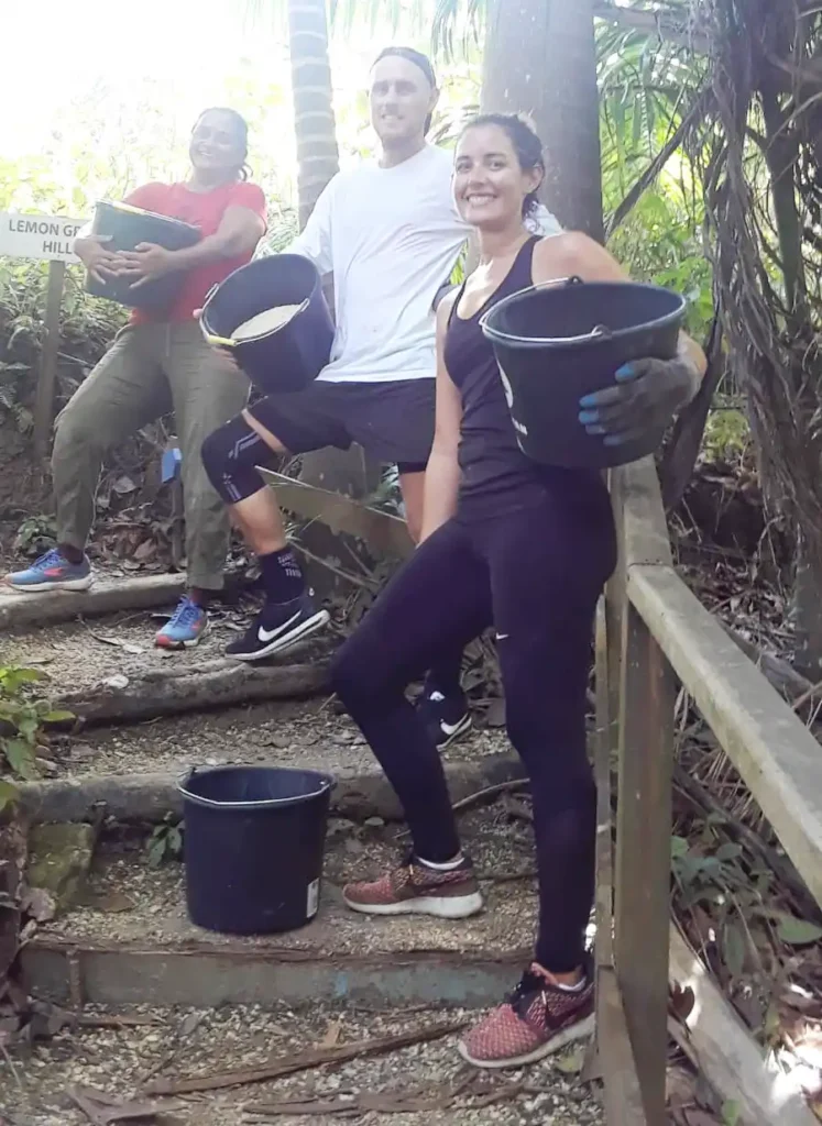 Three volunteers are standing on a wooded trail, each holding a black bucket filled with gravel. The person on the left is wearing a red shirt and olive green pants, the person in the middle is wearing a white shirt and black shorts, and the person on the right is wearing a black tank top and black leggings. They are all smiling and appear to be engaged in a trail maintenance activity, with the background showing lush green foliage and a sign that reads "Lemon Grass Hill."