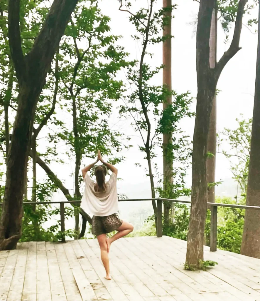 A person is practicing yoga on a wooden deck surrounded by trees. The individual is standing in the tree pose, with one foot placed on the inner thigh of the opposite leg and hands joined above the head. The scene is tranquil, with lush green foliage and a serene, natural backdrop. The deck appears to be elevated, providing a scenic view of the forested area. The person is dressed in a light-colored top and patterned shorts, adding to the relaxed and peaceful ambiance of the setting.