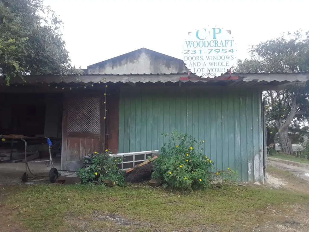 The image shows a small, rustic workshop with a sign on the roof that reads "CP Woodcraft" along with a phone number and a description of services, which include doors, windows, and more. The building has a corrugated metal roof and green, weathered wooden siding. There are some yellow flowers in front of the building, and various tools and materials are scattered around the entrance. The surrounding area includes some trees and a grassy lawn.