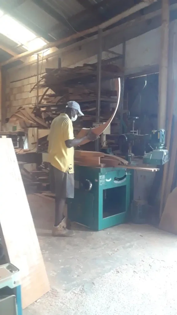 A craftsman in a workshop is carefully examining a piece of wood. He is wearing a yellow shirt, dark shorts, a light-colored cap, and a face mask. The workshop has shelves filled with various wooden planks and tools. The floor is covered with sawdust, and there is a large green woodworking machine in front of him. The workshop has a rustic, industrial feel with exposed brick walls and wooden beams.