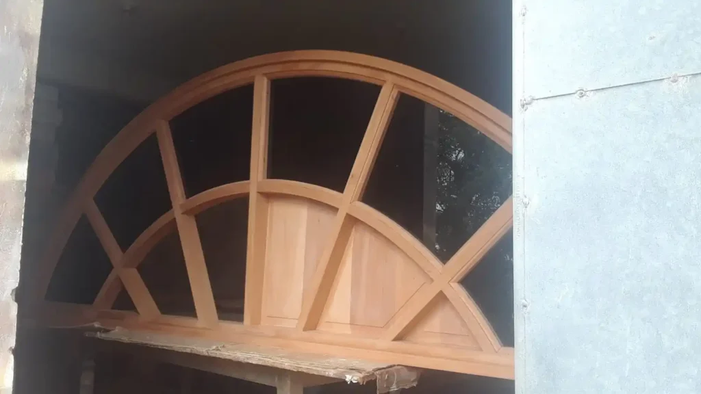 The image depicts a mahogany arch in the process of being constructed. The arch features a series of evenly spaced vertical and horizontal wooden beams, creating a grid-like pattern within the arch. The craftsmanship appears detailed and precise, highlighting the natural beauty and rich color of the mahogany wood. The arch is set within a larger frame or workspace, suggesting it is part of a larger architectural or carpentry project.