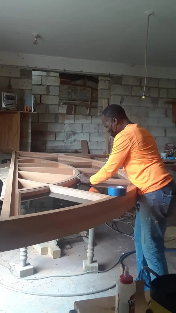 A craftsman wearing a bright orange long-sleeve shirt and jeans is working diligently on a large mahogany arch. The arch appears to be part of a woodworking project for the Barbados parliament buildings. The setting is a workshop with brick walls, various tools, and equipment scattered around. The craftsman is focused on the task, using blue painter's tape to aid in the construction. The arch structure rests on a worktable supported by blocks.