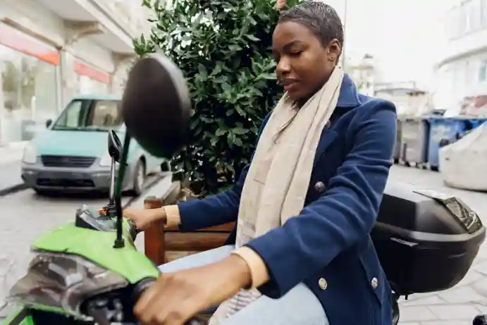 A woman sitting on a green moped, wearing a navy blue coat and a beige scarf. She has short hair and is adjusting the handlebars, looking focused. The scene appears to be on a city street, with a blurred background featuring a parked car, buildings, and some greenery.