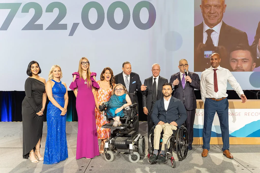 A group photo featuring a diverse group of ten people, including men and women of different ages, ethnicities, and abilities. Two individuals in the front row are in wheelchairs. The group is dressed formally, with women in elegant dresses and men in suits. They are standing on a stage with a large screen behind them displaying the number "722,000". The atmosphere is celebratory, with smiles and a sense of accomplishment evident among the group members.