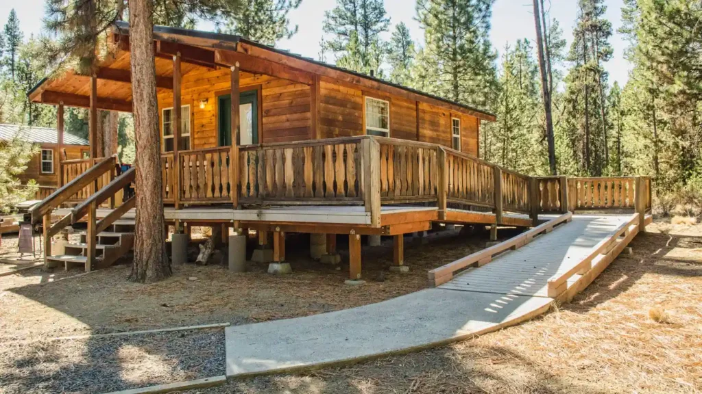 A log cabin surrounded by tall pine trees, featuring a spacious wooden porch with railings. The cabin has a green door and several windows. A wooden ramp and steps provide access to the porch, ensuring wheelchair accessibility. The ground around the cabin is covered with pine needles and some gravel pathways.