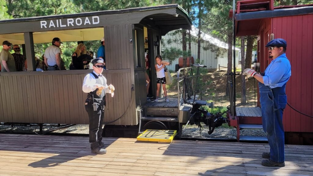 A train conductor and an engineer are standing on a wooden platform next to a stationary train. The conductor, dressed in a white shirt, black vest, and conductor's hat, is holding a pair of gloves. The engineer, wearing blue overalls, a blue shirt, and a flat cap, is holding a tool. The train consists of a brown passenger car with people inside, marked "RAILROAD," and a red caboose. A young girl is stepping off the train, and several passengers can be seen inside the passenger car. The background features trees and a white building.