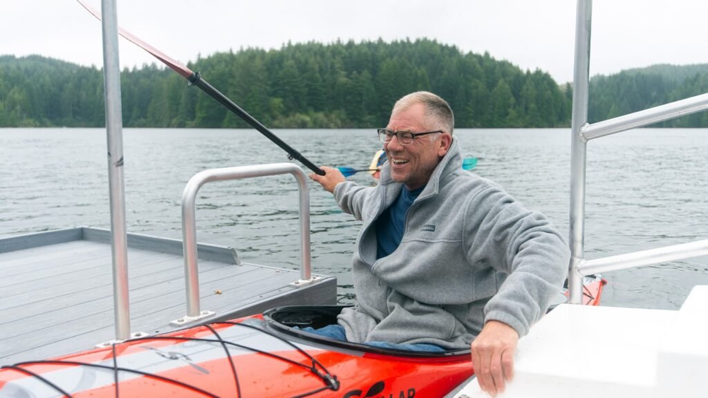 A man smiling while sitting in a kayak on a lake, wearing a gray fleece jacket and glasses. He is holding a paddle and appears to be enjoying the serene, forested surroundings. The weather looks overcast, and there are trees in the background across the water.