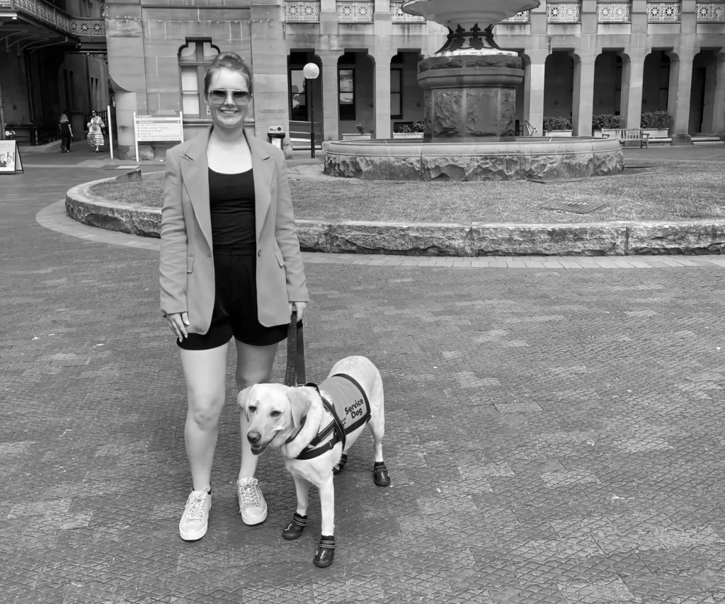 A woman stands outdoors beside a service dog. The woman is wearing a blazer over a top and shorts, with sneakers. She has sunglasses on and her hair is tied back. The service dog, a Labrador Retriever, is wearing a service vest and protective boots on its paws. They are in front of a historic building with arches and a fountain in the background. The scene appears to be in a courtyard with paved ground and a circular grassy area.