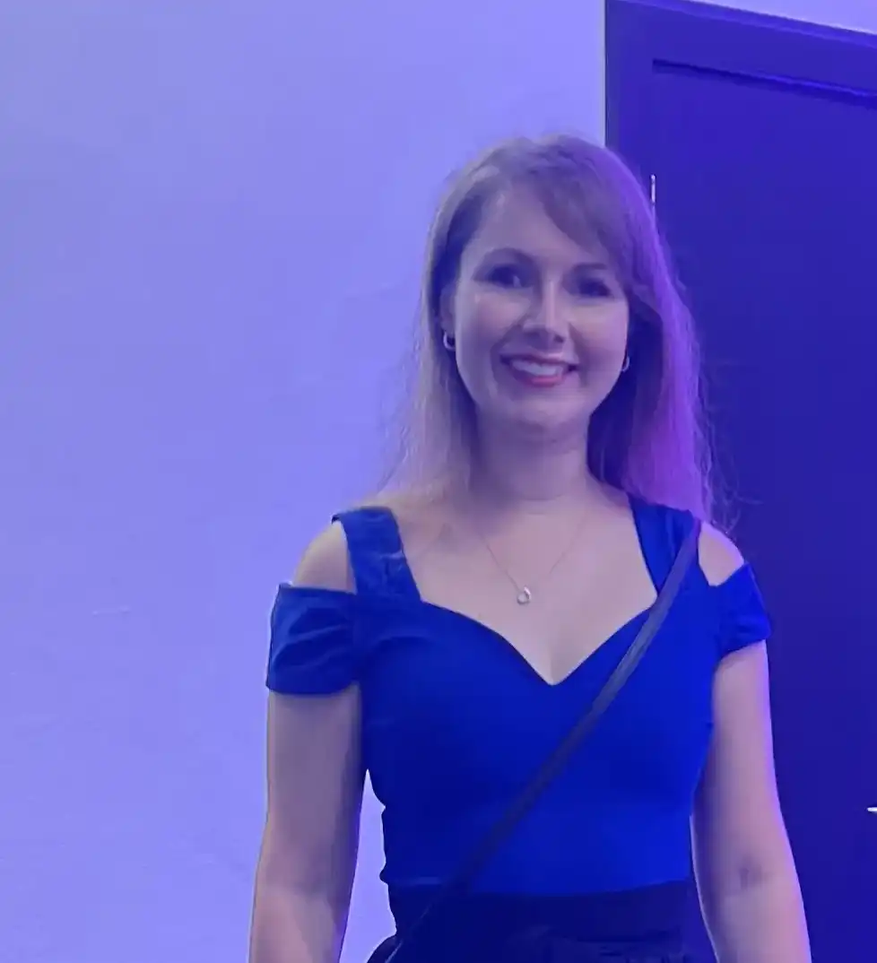 A smiling woman with shoulder-length blonde hair is standing against a light blue and purple background. She is wearing a royal blue dress with cold-shoulder sleeves and a sweetheart neckline. She has a black strap across her body and is wearing a delicate necklace.