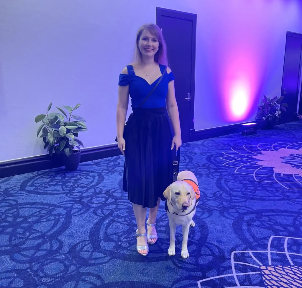 A woman with long brown hair, wearing a blue off-shoulder top and a black skirt, is standing on a carpeted floor with intricate blue patterns. She is smiling and holding the harness of a guide dog, a yellow Labrador wearing an orange vest. The background has a soft purple lighting with potted plants along the walls.