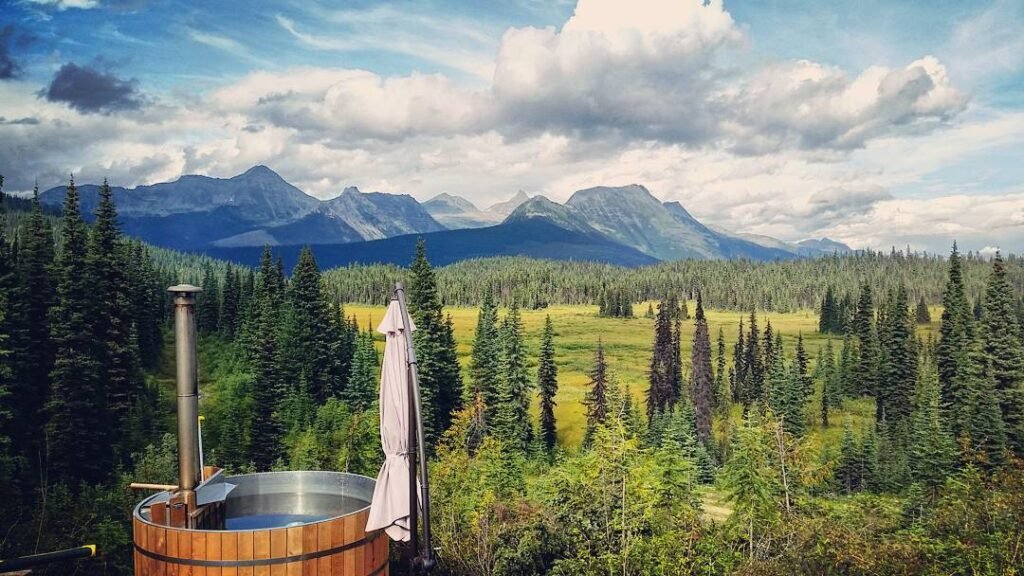 The image depicts a scenic view of a mountainous landscape. In the foreground, there is a round wooden hot tub with a chimney, partially covered by a closed patio umbrella. The middle ground features a lush forest of evergreen trees, which transitions into a vast green meadow. In the background, rugged mountains rise under a partly cloudy sky, creating a picturesque and tranquil outdoor setting.