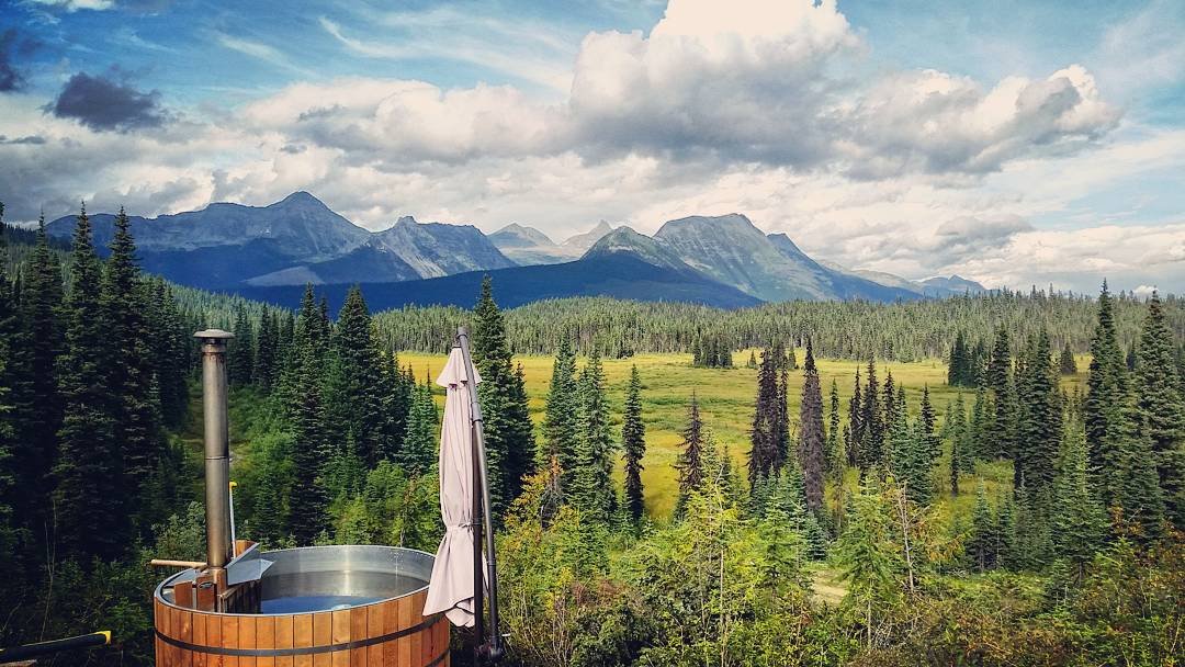 The image depicts a scenic view of a mountainous landscape. In the foreground, there is a round wooden hot tub with a chimney, partially covered by a closed patio umbrella. The middle ground features a lush forest of evergreen trees, which transitions into a vast green meadow. In the background, rugged mountains rise under a partly cloudy sky, creating a picturesque and tranquil outdoor setting.