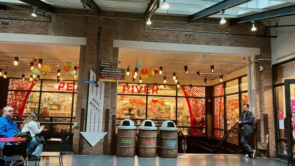 The image depicts an indoor scene, likely within a market or shopping center. There are several elements visible: Storefront: The main focus is on a brightly lit store named "Pearl River" with a red sign. The store has a large glass window displaying various colorful items inside. Lighting: Hanging lights with exposed bulbs add a warm ambiance to the setting. Decoration: Paper lanterns and other decorative items are visible inside and near the store entrance, adding a festive or cultural touch. Signage: There is a directional sign hanging from the ceiling pointing to different stores and amenities, such as Pearl River Mart Foods, ALF Bakery, and more. Additionally, there is a sign indicating the direction to the elevator and restrooms. Seating Area: In the foreground, there are tables and chairs where people are seated. One person in a blue jacket is sitting at a table, and another person is nearby. Trash Bins: Three wooden trash bins are labeled for mixed paper recyclables, trash, and metal/glass/plastic/aluminum cans. Brick Wall: The surrounding walls are made of exposed brick, adding to the industrial aesthetic of the space. People: There are a few people in the scene - one seated at a table, one standing by the wall, and another person partially visible. Overall, the image captures a lively and colorful indoor market scene with a mix of retail and dining elements.