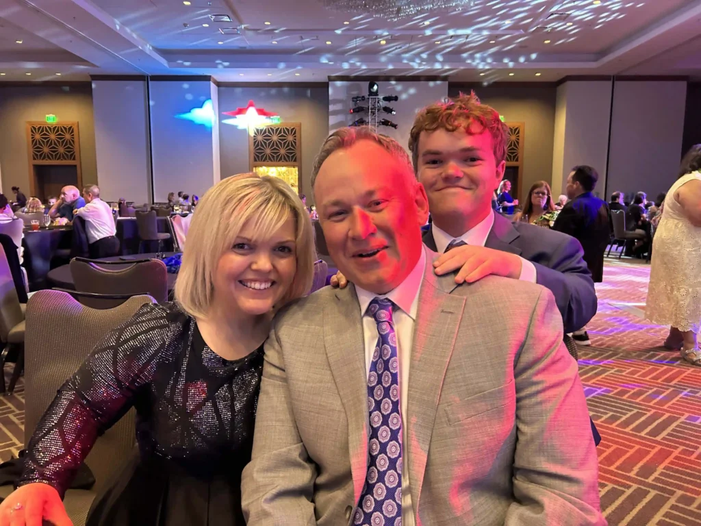 A smiling family is sitting together at a formal event. The woman on the left has blonde hair and is wearing a black, shiny, patterned dress. The man in the center is wearing a light gray suit with a patterned tie, and the young man standing behind them has curly red hair and is wearing a dark suit and tie. They are all posing for the camera, with the young man’s hands resting on the shoulders of the man in the center. The background shows a ballroom with tables, other guests, and colorful lighting.