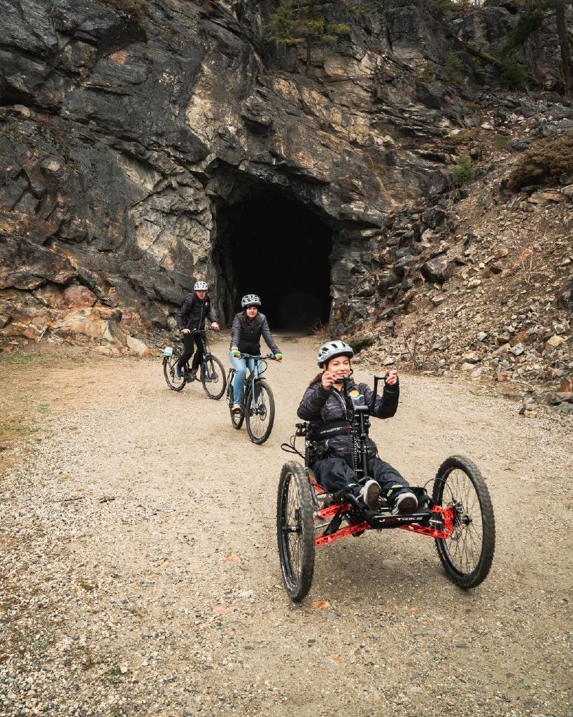 A group of cyclists is exiting a rocky tunnel during a guided bike tour. The first rider is using a red adaptive tricycle, designed for accessibility, and is smiling and gesturing happily. Two other cyclists on standard bicycles follow closely behind, all wearing helmets and casual outdoor attire. The background features rugged rock formations, indicating a mountainous or canyon setting. The path is gravelly, adding to the adventurous atmosphere of the ride.