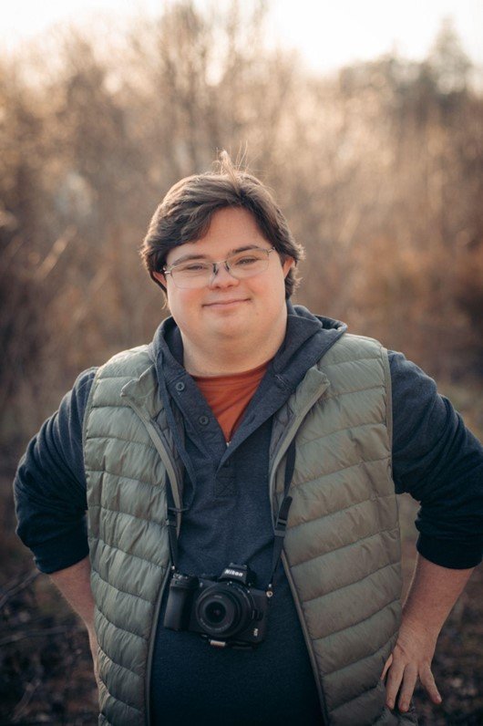 A young man with Down syndrome stands outdoors, smiling at the camera. He has short brown hair and wears glasses. He is dressed in a grey hooded jacket layered with a green quilted vest. A camera is hanging from a strap around his neck. The background is a blurry natural setting with leafless trees, suggesting it might be fall or winter. The light is soft, creating a warm and pleasant atmosphere.