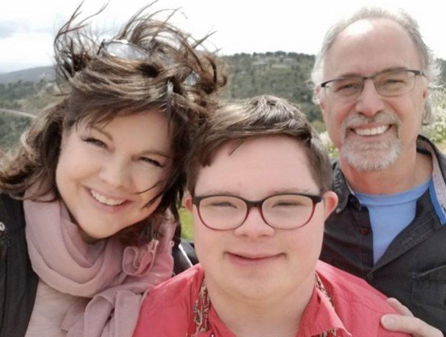 A cheerful family photo featuring three people outdoors on a windy day. On the left, a woman with shoulder-length brown hair, wearing a light pink scarf, smiles brightly. In the middle, a young man with short brown hair and glasses, wearing a red shirt, smiles warmly. On the right, a man with short gray hair, a beard, and glasses, dressed in a dark shirt with a blue collar, smiles broadly. The background shows a scenic landscape with greenery and hills.