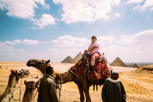 A person is riding a camel adorned with a colorful, patterned saddle blanket in the desert with pyramids visible in the background. The sky is partly cloudy. Two local guides stand near the camel, guiding it. The scene suggests a tourist experience in Egypt, likely near the Giza pyramids.