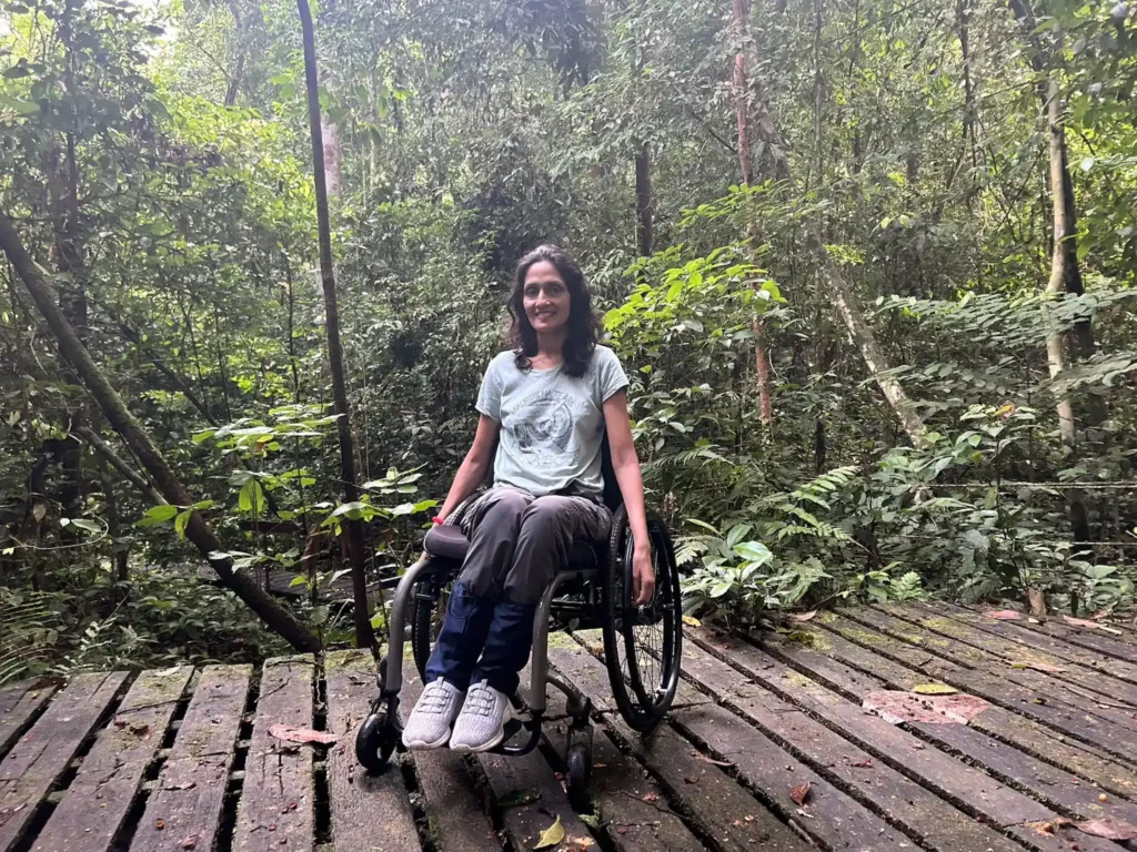 A woman in a wheelchair is on a wooden boardwalk surrounded by lush, green rainforest. She is wearing a light green T-shirt, blue pants, and white sneakers. She has dark hair and is smiling at the camera. The scene is serene, with dense foliage and trees in the background, indicating a natural and peaceful environment.