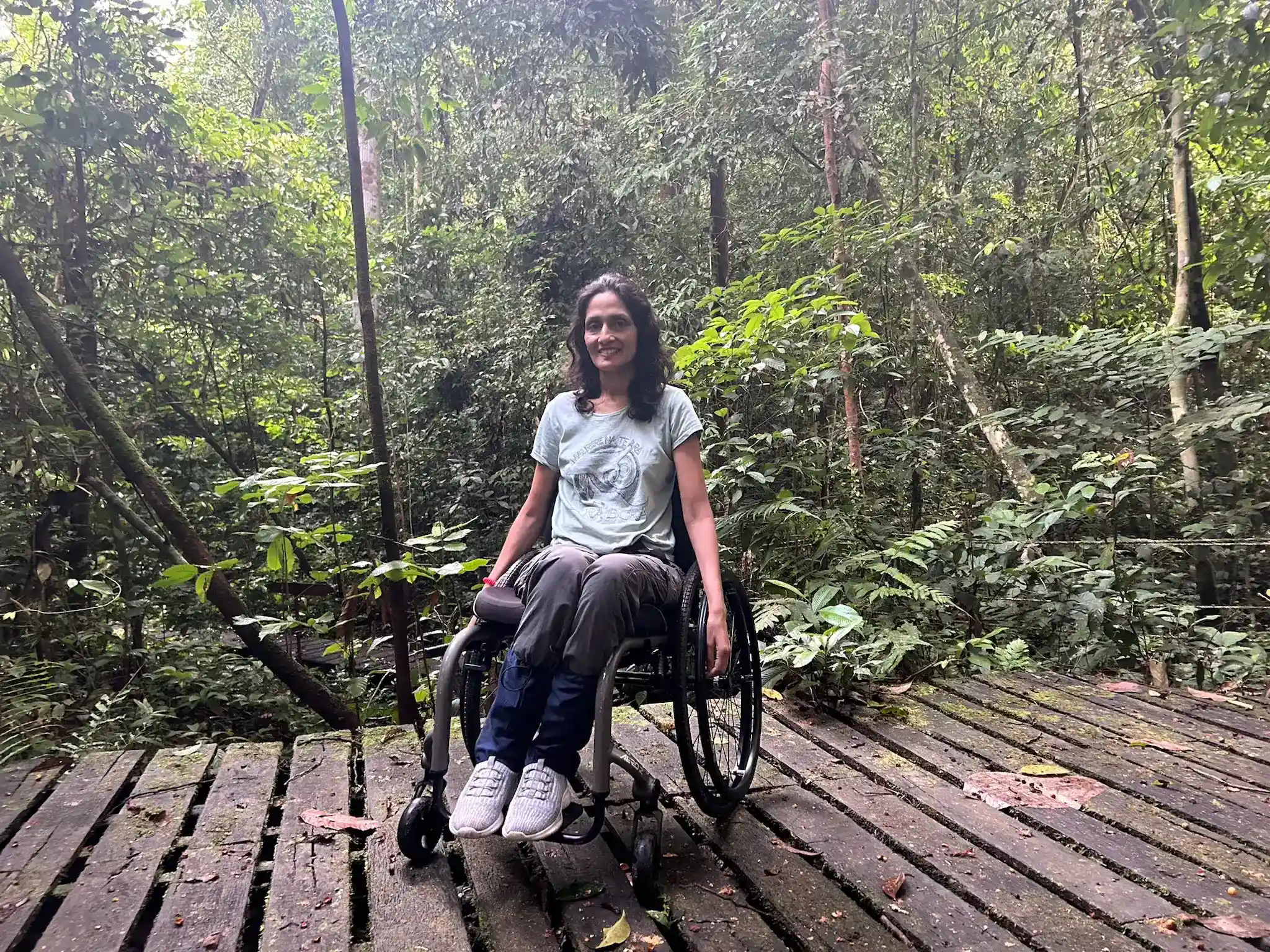 A woman in a wheelchair is on a wooden boardwalk surrounded by lush, green rainforest. She is wearing a light green T-shirt, blue pants, and white sneakers. She has dark hair and is smiling at the camera. The scene is serene, with dense foliage and trees in the background, indicating a natural and peaceful environment.