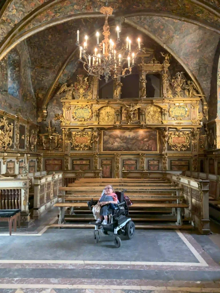 A person in a wheelchair is sitting in the center of an opulent, historic church interior. The church features intricate gold detailing, ornate woodwork, and a large, elaborate chandelier hanging from a vaulted ceiling decorated with rich, colorful frescoes. The wooden pews are arranged symmetrically, and the overall atmosphere exudes grandeur and antiquity.