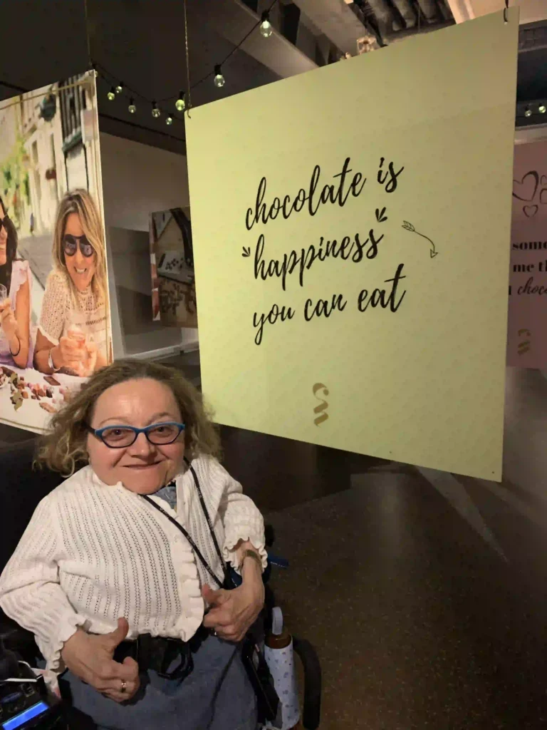 A woman is standing in front of a sign at the Chocolate Nation museum. The sign reads, "Chocolate is happiness you can eat." The woman is smiling, giving two thumbs up, and wearing a white, knitted cardigan over a blue dress. She has glasses with blue frames and curly hair. The background features another poster with two women enjoying ice cream. The setting appears to be a museum exhibit dedicated to chocolate.