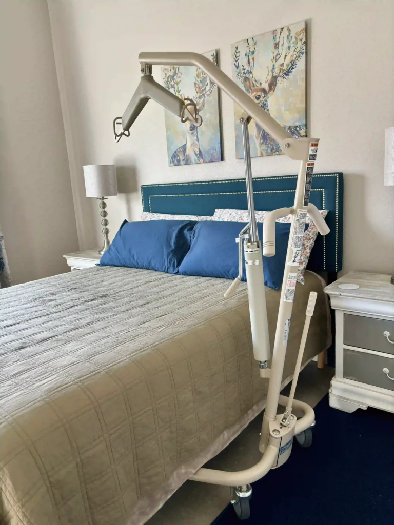Invacare Manual Hydraulic Lift : The image shows a bedroom with a bed that has a teal headboard and blue pillows. There are two nightstands on either side of the bed, each with a lamp on it. Above the bed, there are two pieces of artwork depicting deer with floral designs. In front of the bed, there is an Invacare Manual Hydraulic Lift, typically used to assist in lifting and moving individuals with mobility challenges. The lift has a metal frame with wheels for easy mobility and a large arm that extends over the bed. The room appears well-decorated and comfortable, combining functionality with aesthetics.