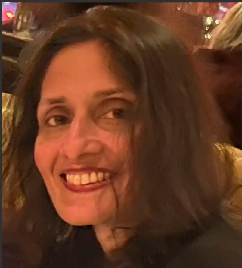 A woman with long dark hair is smiling warmly. She has a relaxed expression and is in a dimly lit setting, possibly a restaurant or a similar cozy environment. The background is blurred, highlighting her face and her pleasant smile.