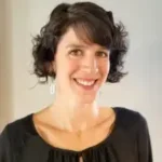 A smiling woman with short, curly dark hair wearing a black top and white earrings, posing against a plain background.
