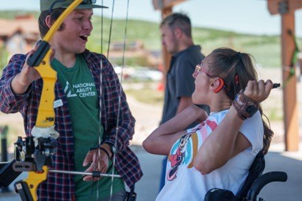 A woman in a wheelchair is practicing archery with assistance. She is wearing a white T-shirt with a colorful design and glasses. The instructor beside her is holding a yellow bow, wearing a green shirt with the "National Ability Center" logo, a plaid shirt over it, and a cap. They are both smiling and engaged in the activity, with a background showing an outdoor setting with another person in the distance.