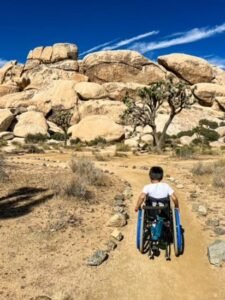 A little boy in a wheelchair is on a dirt trail in a desert landscape, with large boulders and Joshua trees in the background. The sky is clear and blue with a few wispy clouds. The trail is marked by small rocks lining its edges.